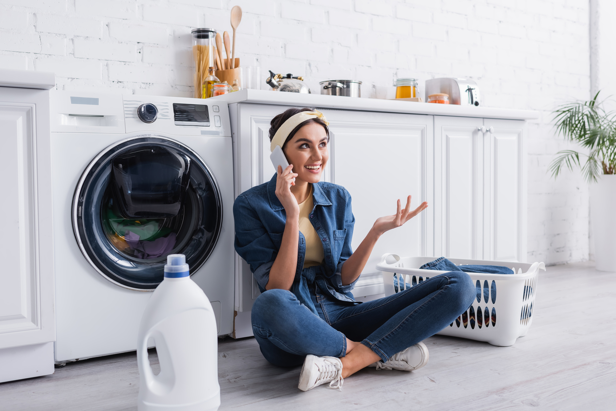 Smiling housewife talking on smartphone near detergent and washing machine in kitchen.jpg